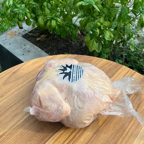 A whole broiler chicken in vacuum packaging, displayed on a wooden surface with foliage in the background.