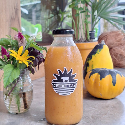 Bottle of smoothie with 'The Good Food Harvest' label on a table with flowers and squash.